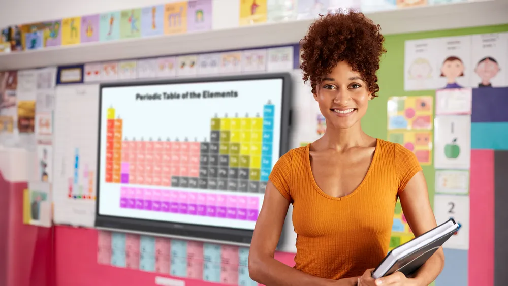 a teacher standing in a classroom. a viewboard is visible on the wall behind her with the periodic table of elements displayed.