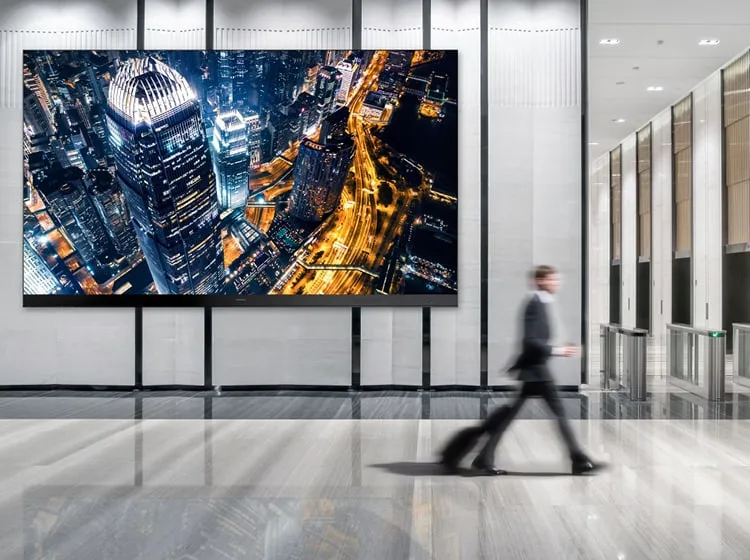 A business man with a suitcase walks speedily past a large display in a public lobby that is showing a cityscape